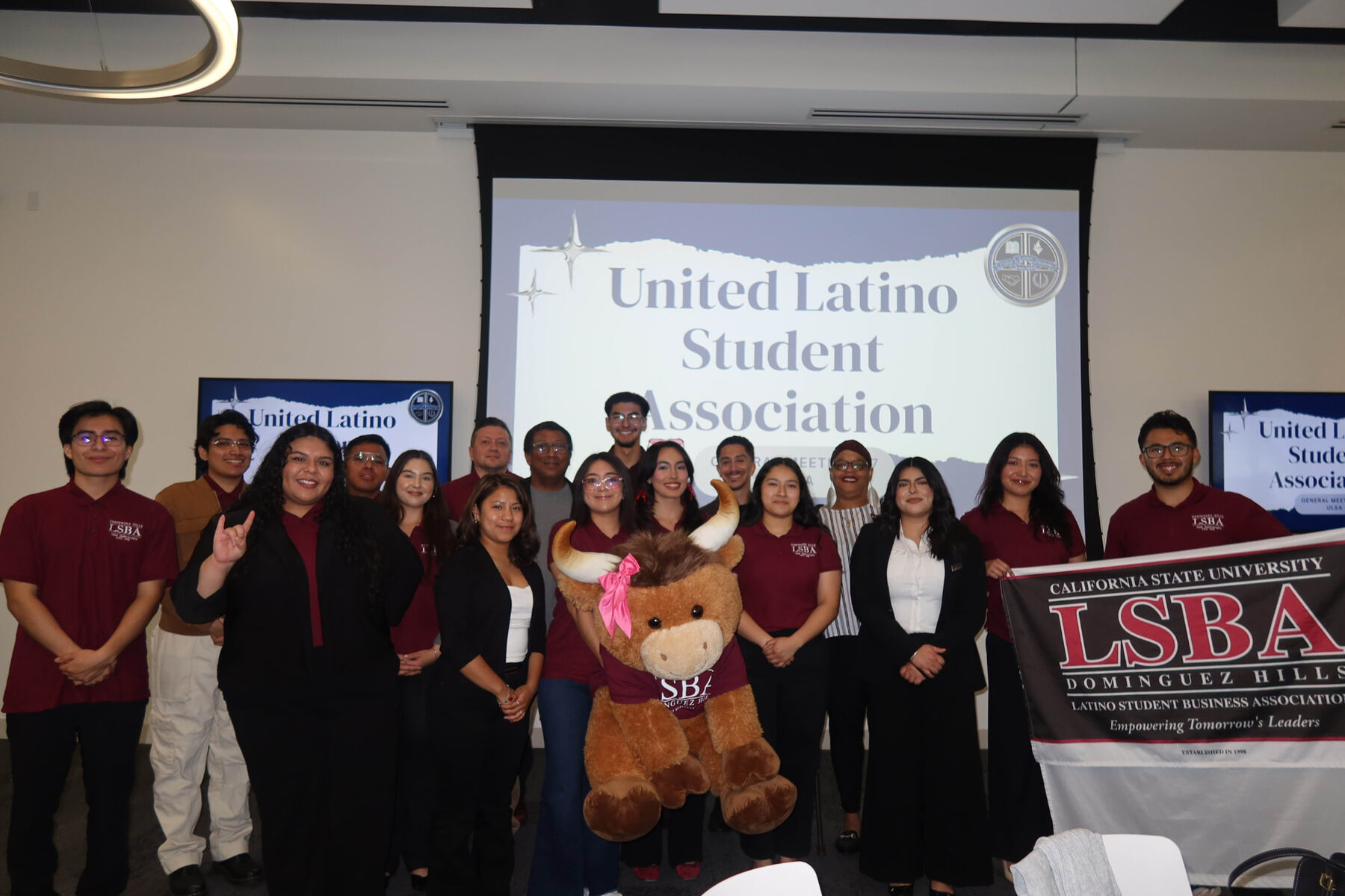 LSBA members in maroon polos posing with the CSUDH LSBA banner and mascot at a United Latino Student Association general meeting.